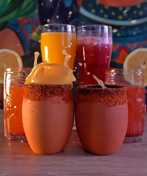 Vasos de barro con borde escarchado y vasos altos con jugos de naranja y mora en pleno movimiento, con salpicaduras, mural de fondo.