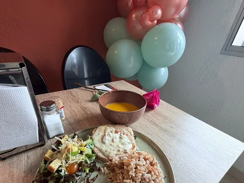 Almuerzo servido en una mesa junto a la pared terracota: plato con pollo en salsa, arroz y ensalada, una sopa y una rosa, con un racimo de globos verde menta y coral al fondo.