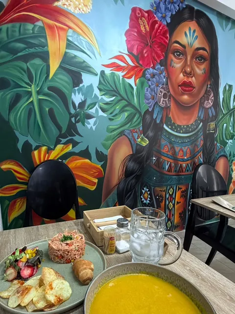 Almuerzo servido frente al mural del salón: un plato con un molde de arroz con maíz, ensalada con fresas, papas en rodajas y un pan, una sopa crema amarilla y un vaso con hielo; al fondo, la mujer indígena pintada entre hojas y flores tropicales.