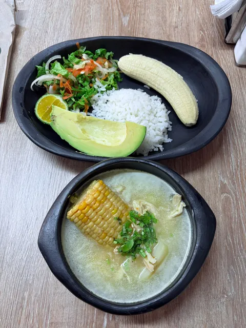 Plato con arroz, aguacate, plátano y ensalada, y una sopa con mazorca, pollo desmechado y cilantro, en la mesa.