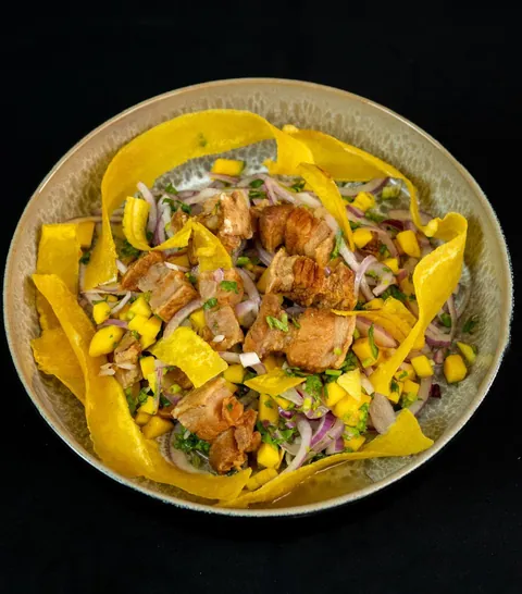 Bowl con trozos de cerdo dorado, mango en cubos, cebolla morada, cilantro y láminas de plátano frito, sobre fondo negro.