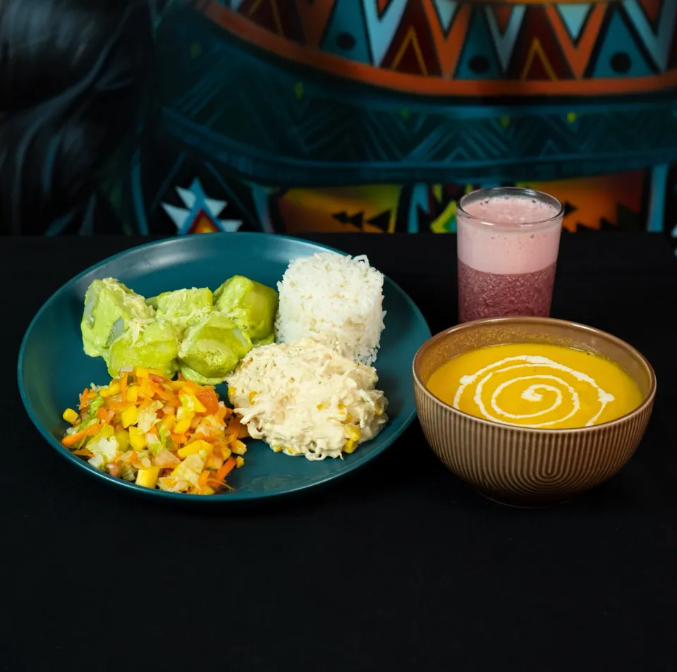 Almuerzo de estudio: plato con trozos gratinados en salsa verde, arroz blanco, desmechado en salsa blanca con maíz y ensalada picada, junto a una sopa crema con un espiral de crema y un jugo, sobre fondo negro con el mural detrás.