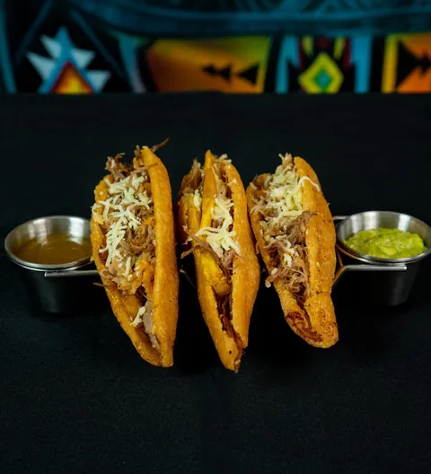 Tres tacos de maíz dorados rellenos de carne desmechada y queso, servidos de pie con una salsa marrón y un guacamole a los lados, sobre fondo negro.