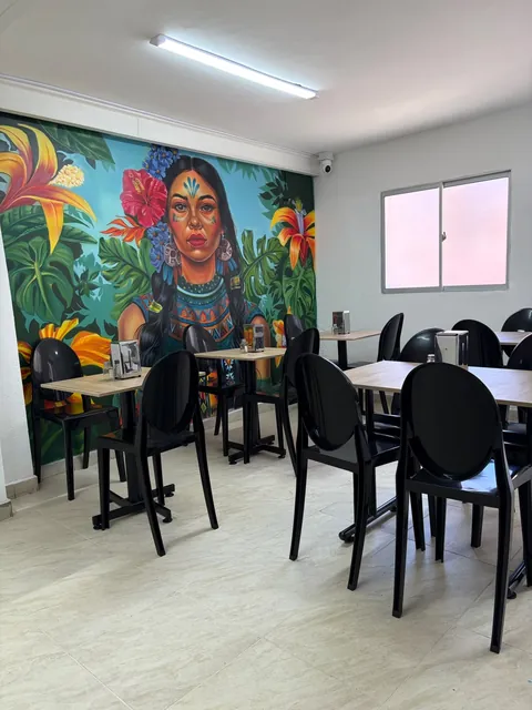 Salón de Resplandor Restaurante con el mural pintado de una mujer indígena entre flores y hojas tropicales, y mesas con sillas negras alrededor.