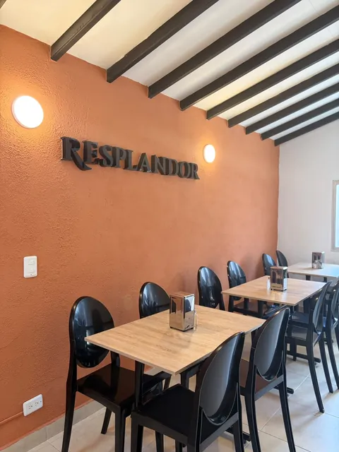 Salón de Resplandor Restaurante: pared terracota con las letras oscuras en relieve «RESPLANDOR» y dos lámparas redondas, vigas negras en el techo blanco, mesas de madera clara y sillas negras.